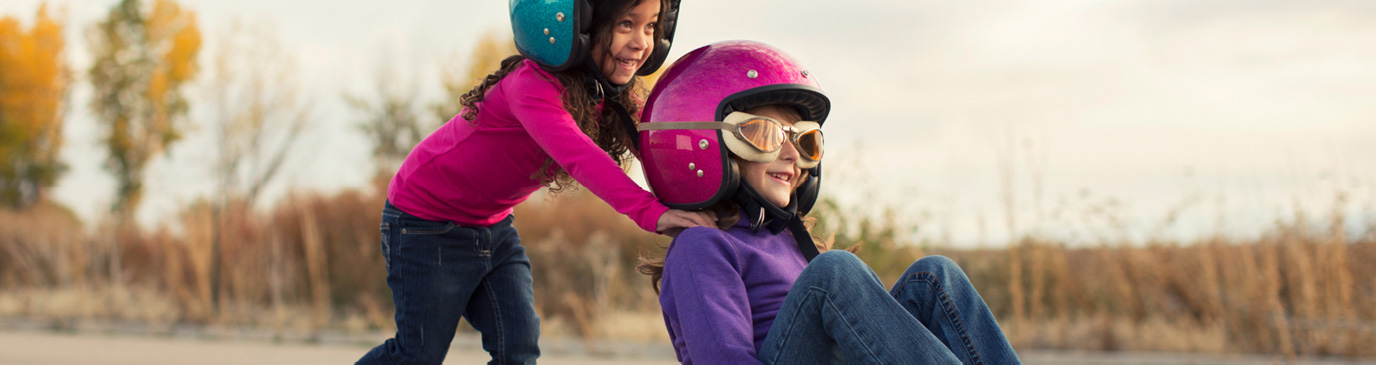 Two smiling girls wearing helmets, one sat on skateboard wearing goggles, being pushed by the other